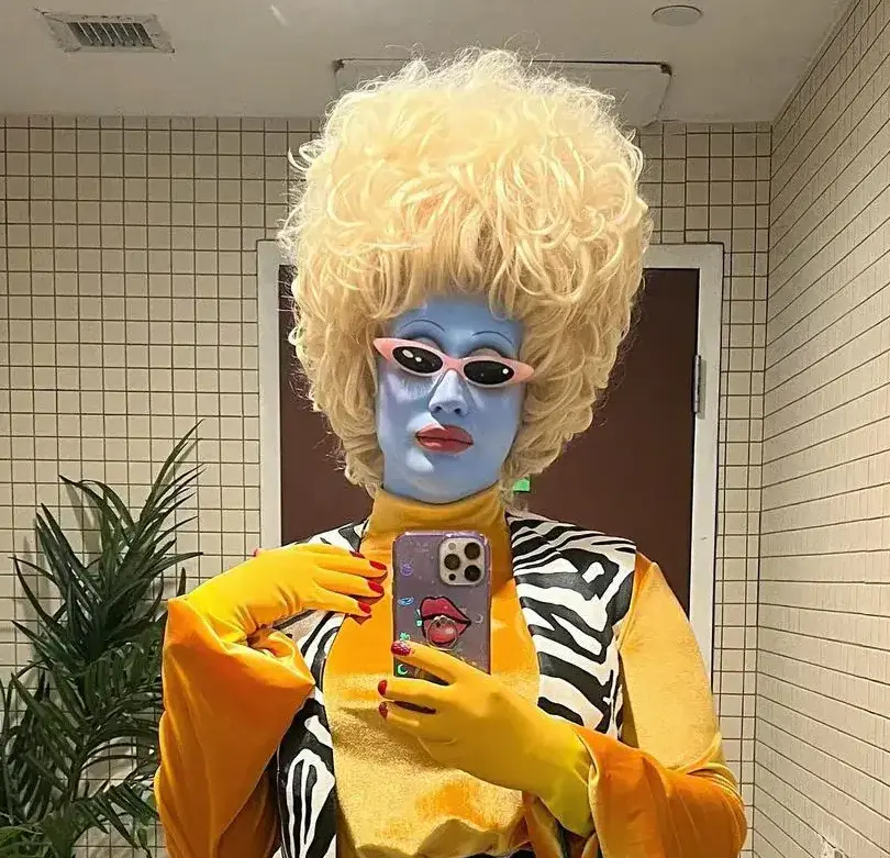 Juno Birch in signature blue alien makeup with yellow hair and retro sunglasses performing drag queen character